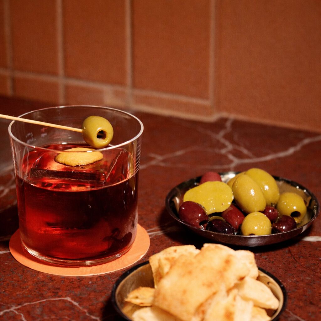 Un vaso de bebida roja con una aceituna en brocheta, un cuenco de aceitunas y un plato de pan plano sobre una encimera de mármol.