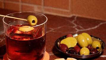 Un vaso de bebida roja con una aceituna en brocheta, un cuenco de aceitunas y un plato de pan plano sobre una encimera de mármol.
