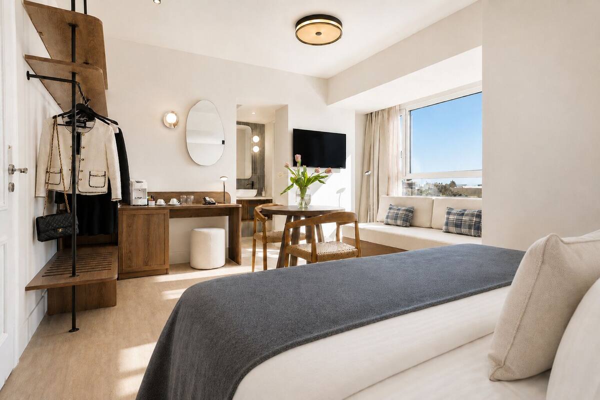 Une chambre d’hôtel moderne équipée d’un lit, d’un bureau, d’une chaise, d’une petite table ornée d’un vase de fleurs, d’un miroir et d’une grande fenêtre.