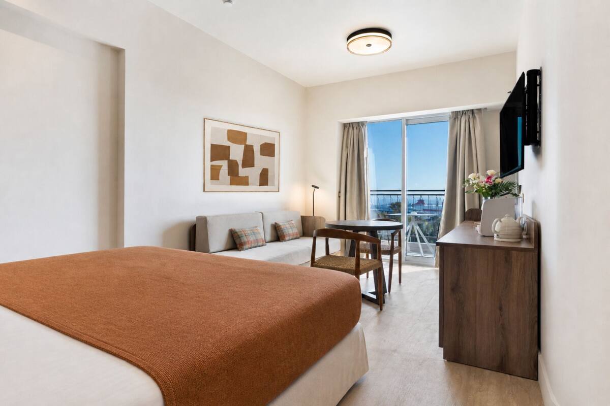 Une chambre d’hôtel avec un lit, un canapé, une petite table, une chaise, un bureau, une télévision et un balcon avec vue.