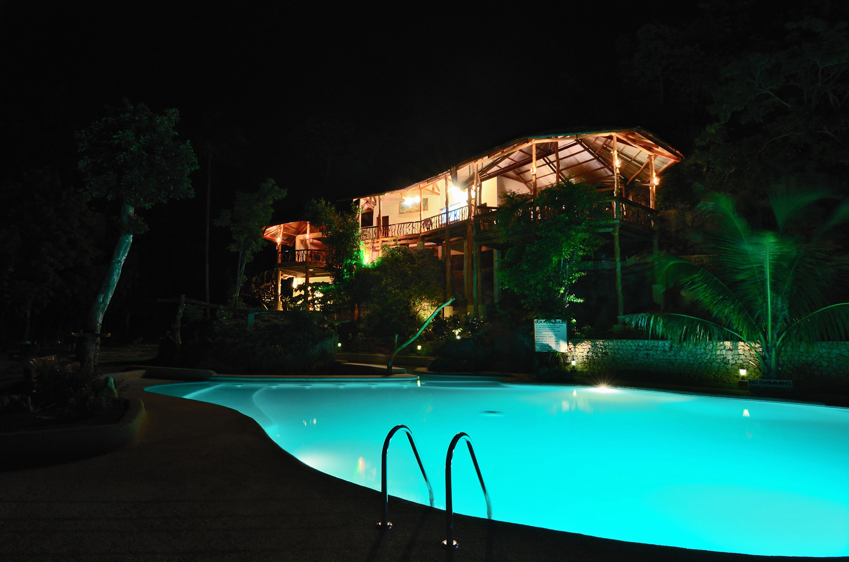 A nighttime view of a resort with a swimming pool, a well-lit building, and surrounding greenery.