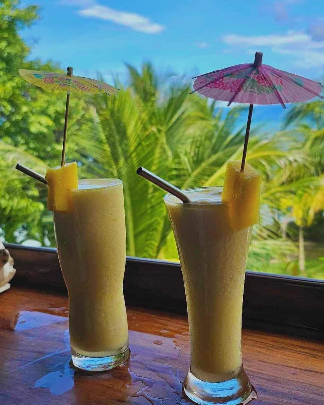 Two glasses of a beverage with pineapple garnishes and umbrellas, placed on a wooden surface with a view of palm trees.