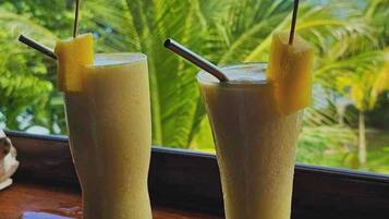 Two glasses of a beverage with pineapple garnishes and umbrellas, placed on a wooden surface with a view of palm trees.