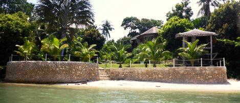 A coastal resort with a stone wall, palm trees, and a thatched-roof structure.