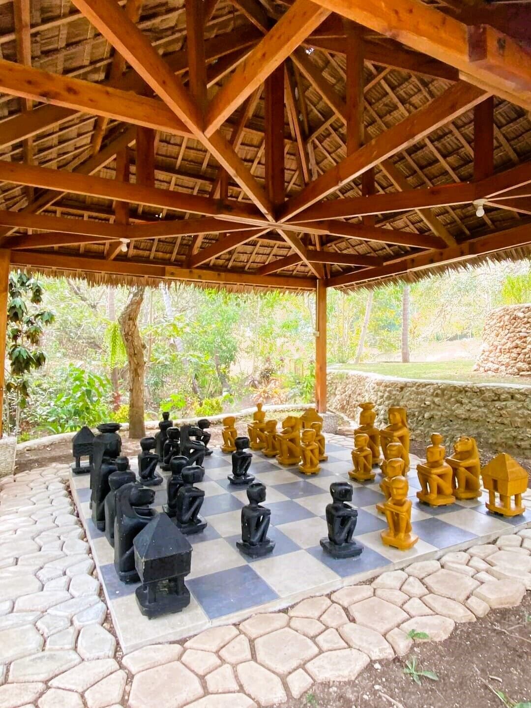 A chessboard set up outdoors under a wooden pavilion with a thatched roof, surrounded by greenery.