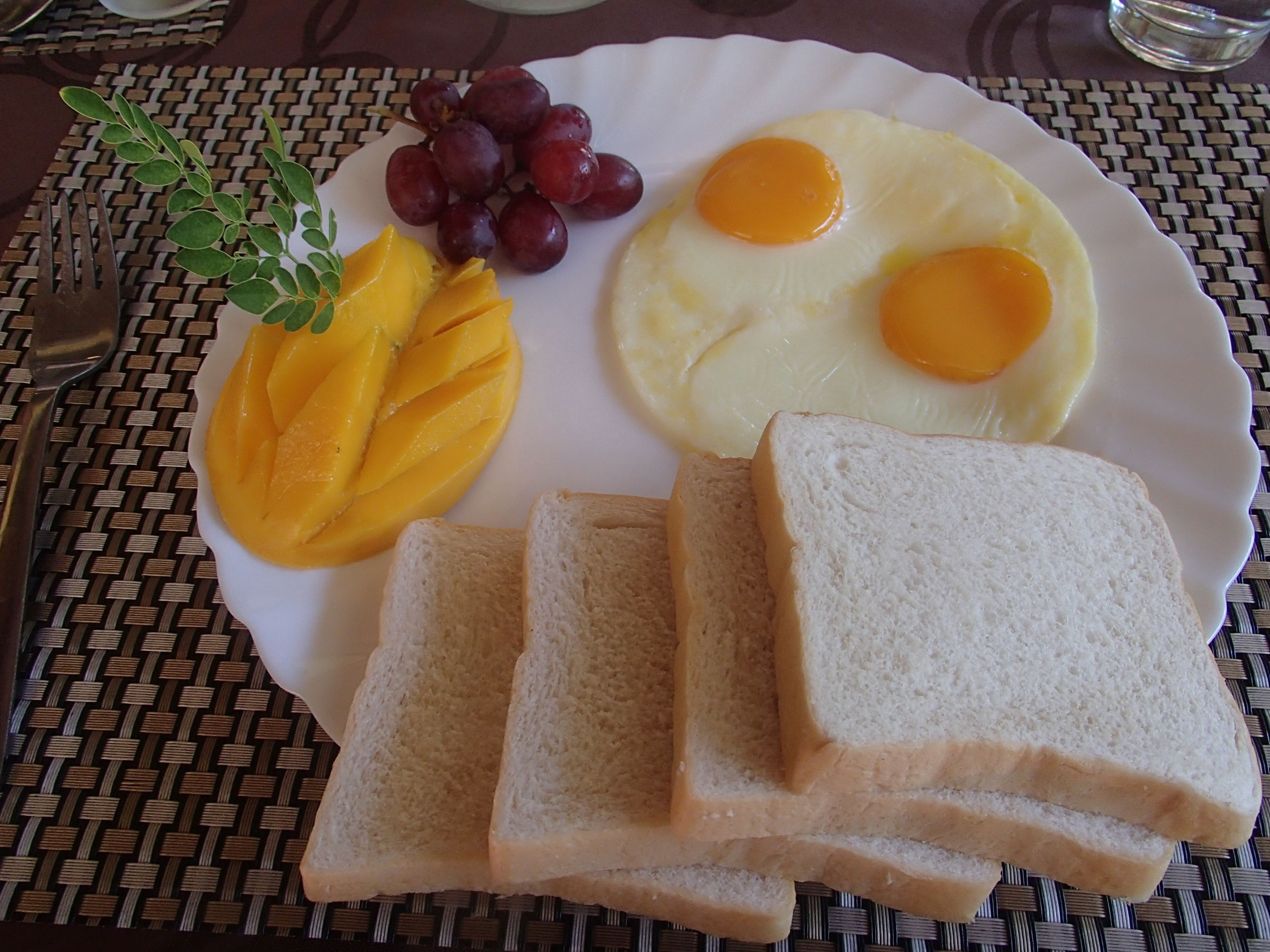 A plate with eggs, mango slices, grapes, and white bread on a woven tablecloth.