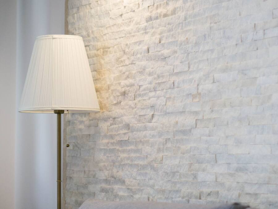 A lamp with a white shade and a wall-mounted light fixture against a textured brick wall.