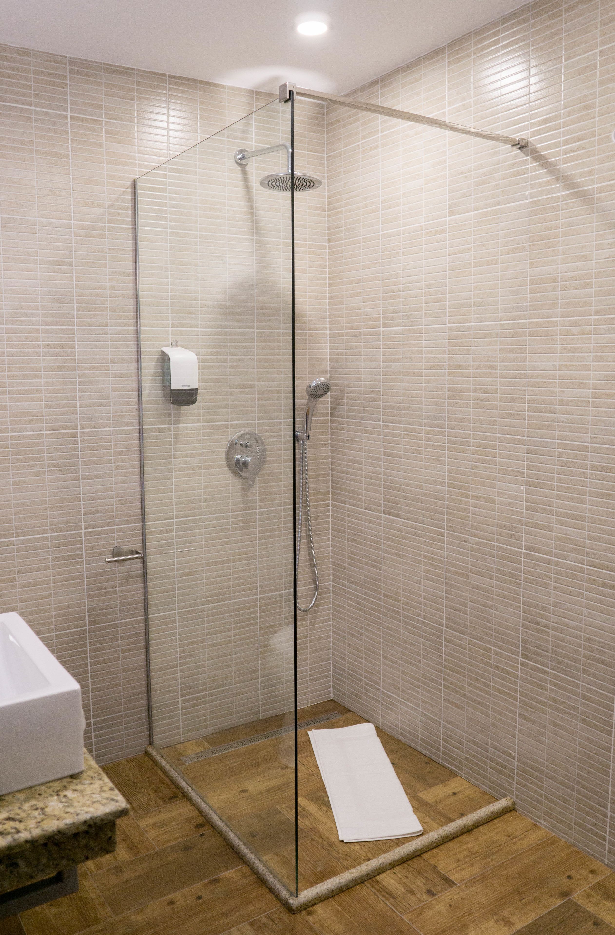 A glass-enclosed shower with beige tiles, a wooden floor, and a white towel on the floor.
