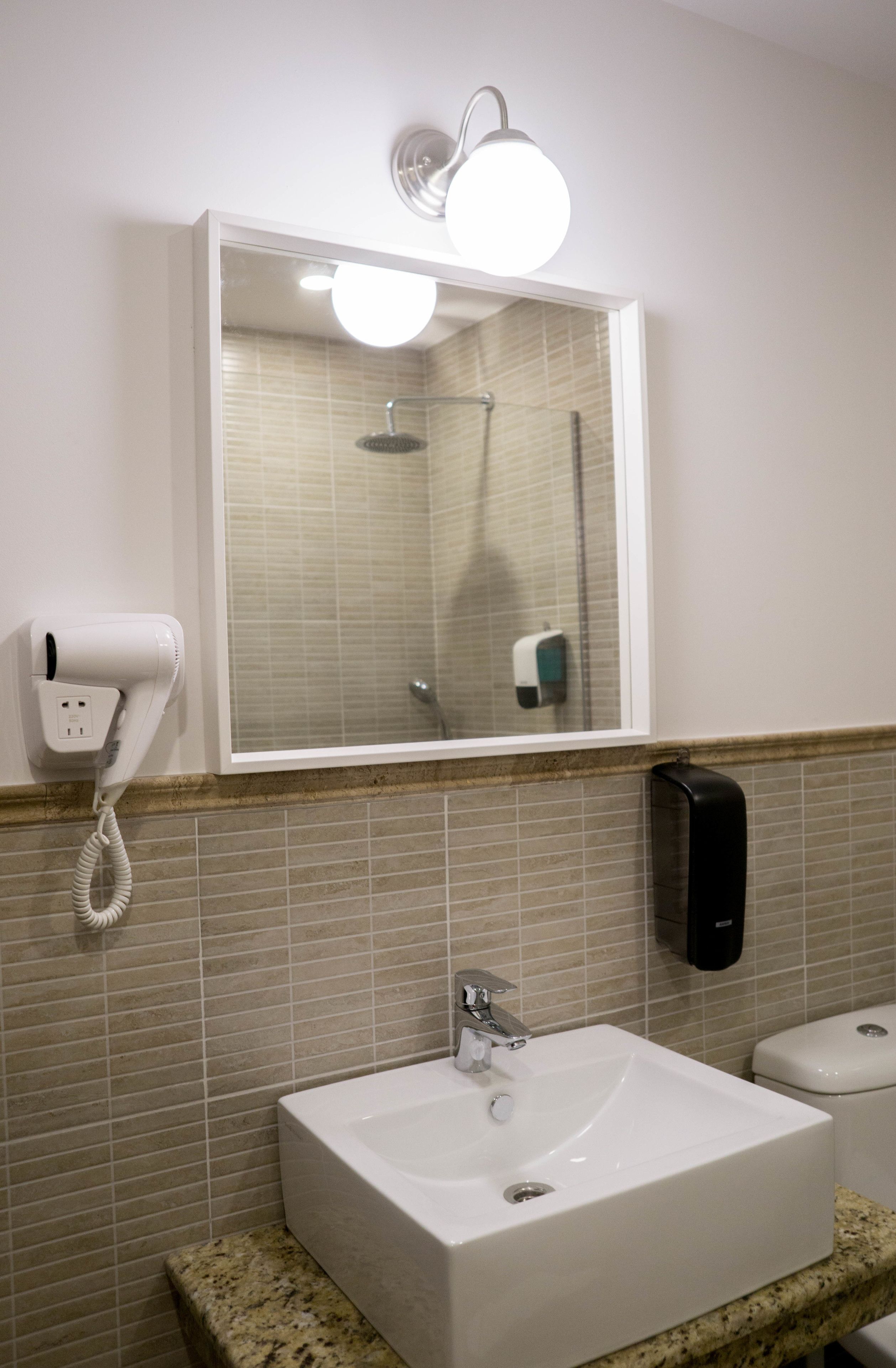 A bathroom with a white sink, a mirror above it, a hairdryer on the wall, and a hand dryer.