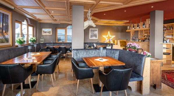 A restaurant with wooden tables and black leather chairs, a bar area with shelves of bottles, and a star decoration on the wall. - Hotel Somont (Selva di Val Gardena)