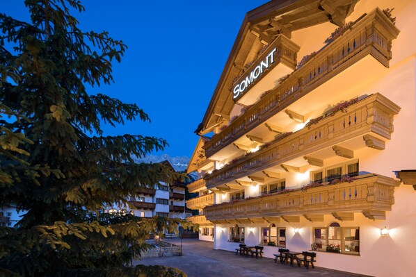 A multi-story hotel with balconies adorned with flowers, a well-lit facade, and a clear sky. - Hotel Somont (Selva di Val Gardena)