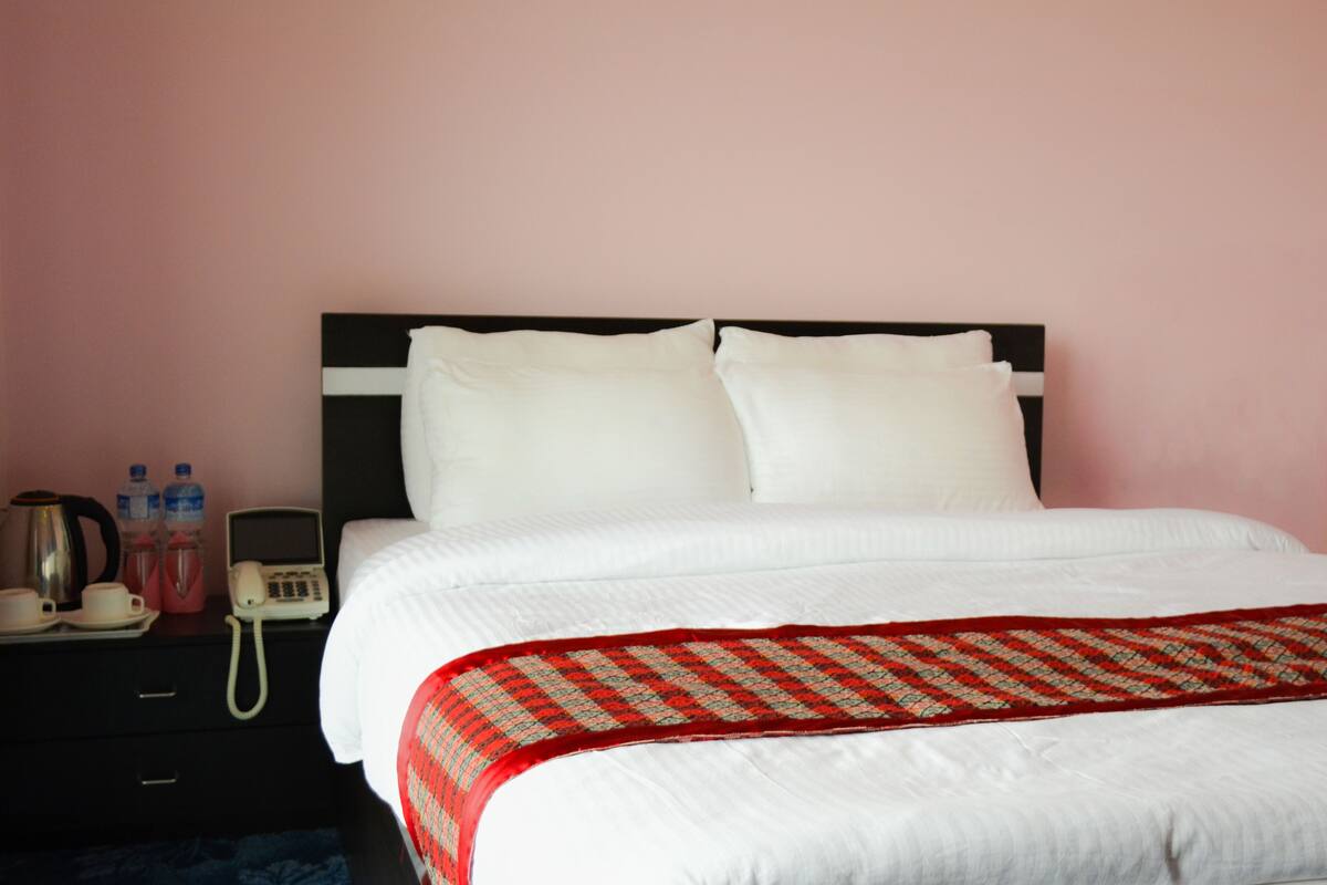 Una cama bien hecha con ropa blanca y un edredón de color rojo y verde. Se observa un mesita de noche negra con un teléfono blanco, una tetera negra y dos botellas de agua. La pared es de color rosa.