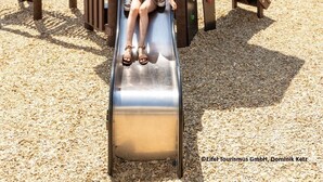 A playground with a slide, surrounded by a wooden structure and a stone building with towers in the background. - Seehotel Maria Laach (Glees)