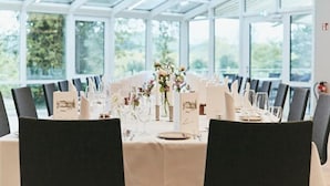 A dining area with a long table set for a meal, surrounded by chairs, with a glass ceiling and large windows. - Seehotel Maria Laach (Glees)