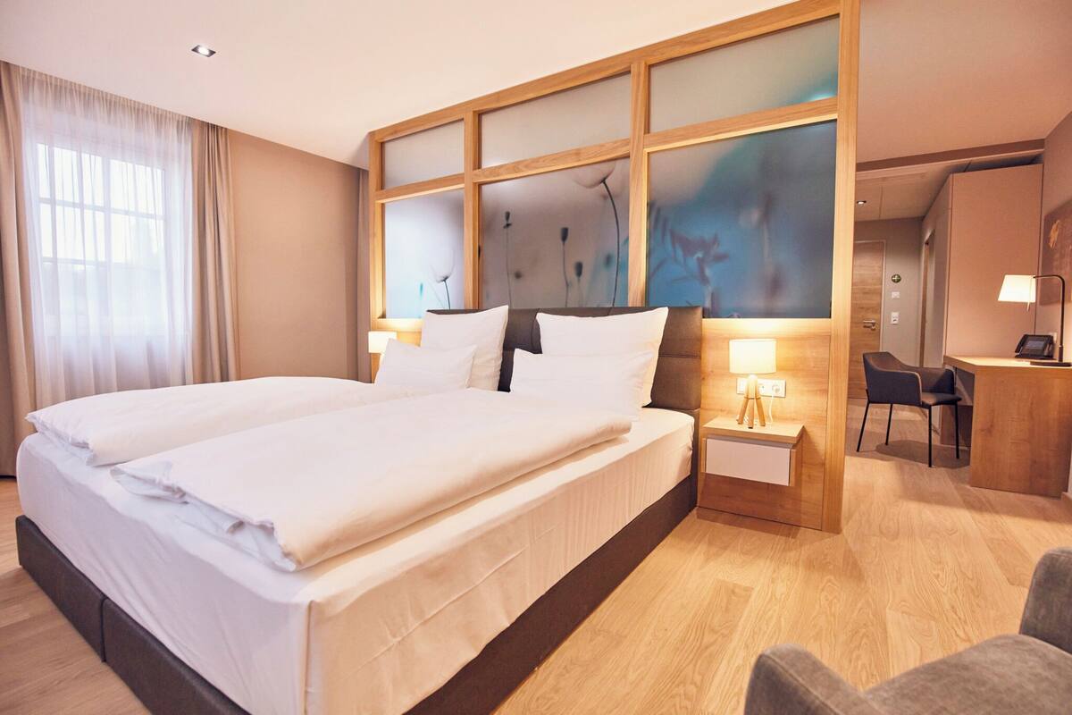 Ein Hotelzimmer mit einem großen Bett, einem Schreibtisch mit Stuhl und einem großen Fenster mit Vorhängen.