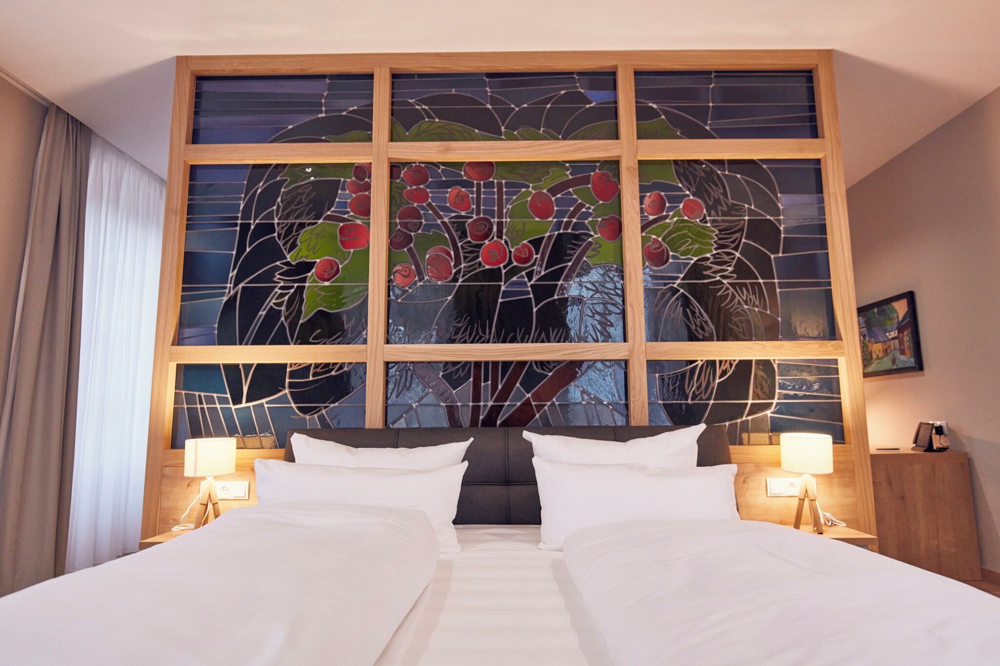 Ein Hotelzimmer mit großem Fenster, das ein Buntglasdesign zeigt, zwei Betten, Nachttischen mit Lampen und einem gerahmten Bild an der Wand.