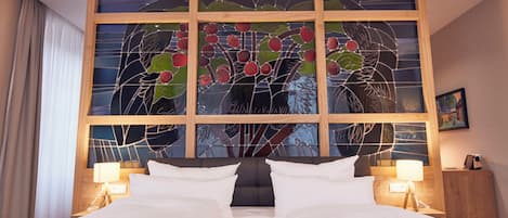 Ein Hotelzimmer mit großem Fenster, das ein Buntglasdesign zeigt, zwei Betten, Nachttischen mit Lampen und einem gerahmten Bild an der Wand.