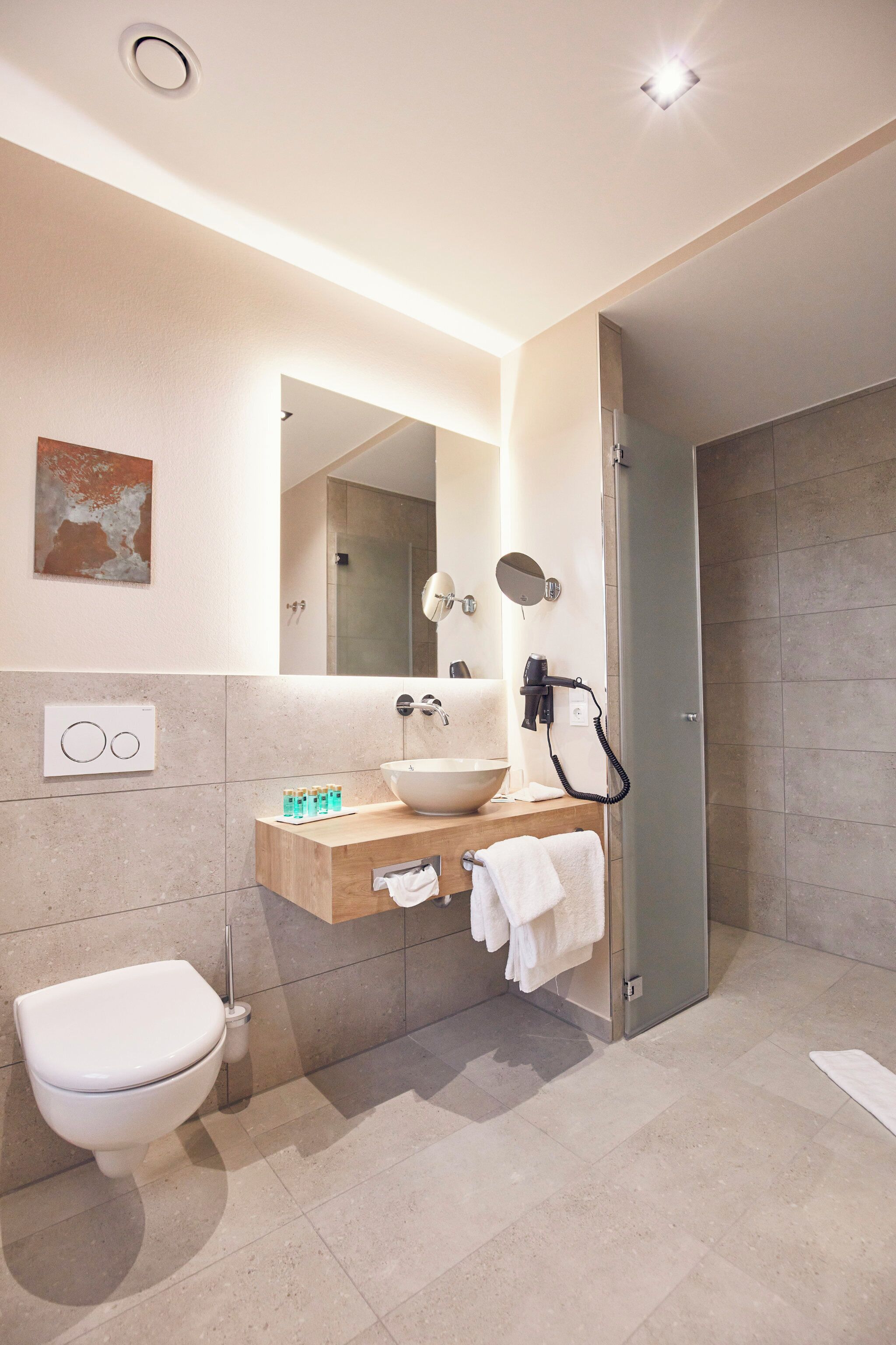 A modern bathroom with a white toilet, a wooden vanity with a sink, a mirror, and a shower area.