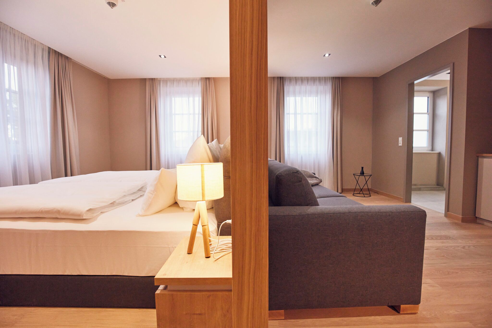 A modern hotel room with a large bed, a sofa, and a wooden nightstand with a lamp.