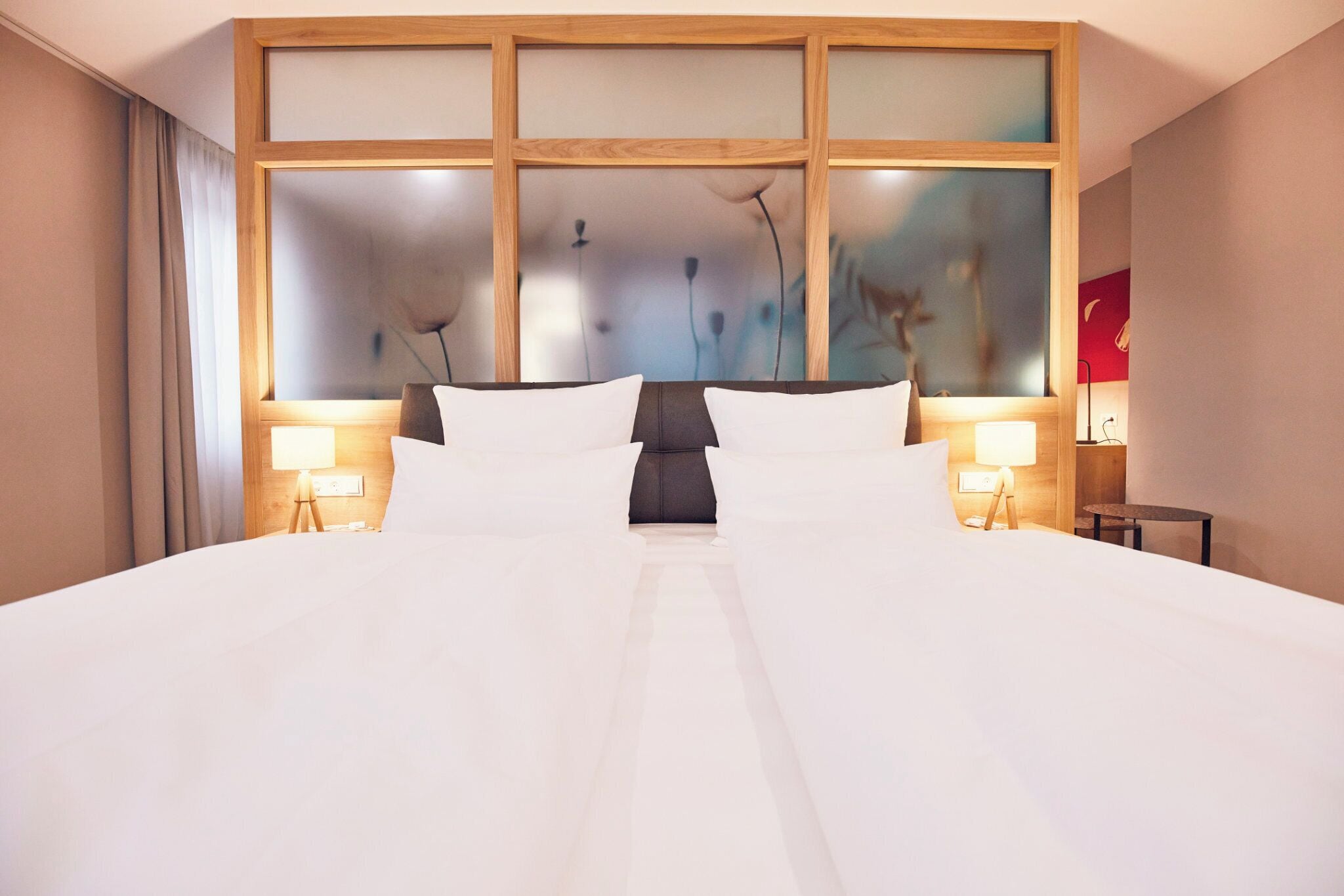 A hotel room with a large bed, two bedside lamps, a wooden headboard with frosted panels, and a large window with a view of trees.