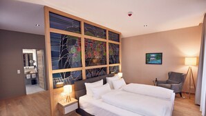 A hotel room with a large bed, a wooden headboard with stained glass, a bedside table with a lamp, a gray armchair, and a painting on the wall. - Seehotel Maria Laach (Glees)