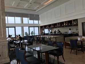 A spacious dining area with multiple tables and chairs, a kitchen in the background, and large windows providing a view. - Luxury Apartment Ocean View (Da Nang)