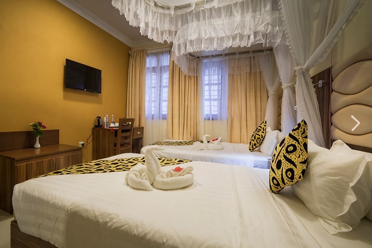 Ein Hotelzimmer mit zwei Betten, einem an der Wand montierten Fernseher, einem hölzernen Schminktisch und einem Fenster mit Vorhängen.
