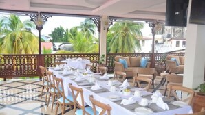 Un espacio al aire libre cubierto para comer, con mesas preparadas para una comida, rodeado de mobiliario de mimbre y palmeras al fondo.