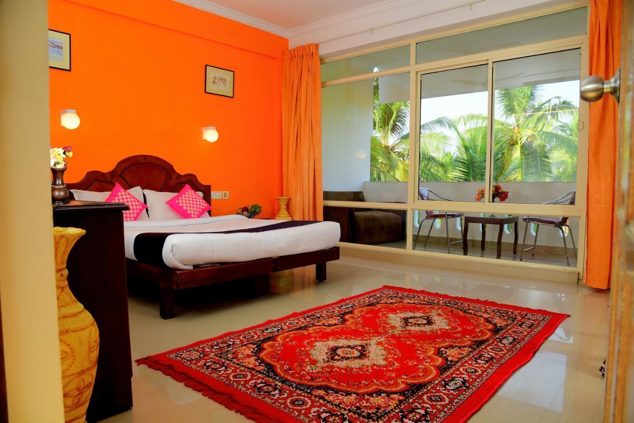 A hotel room with a large bed, a red patterned rug, a desk with a chair, and a large window with a view of palm trees.