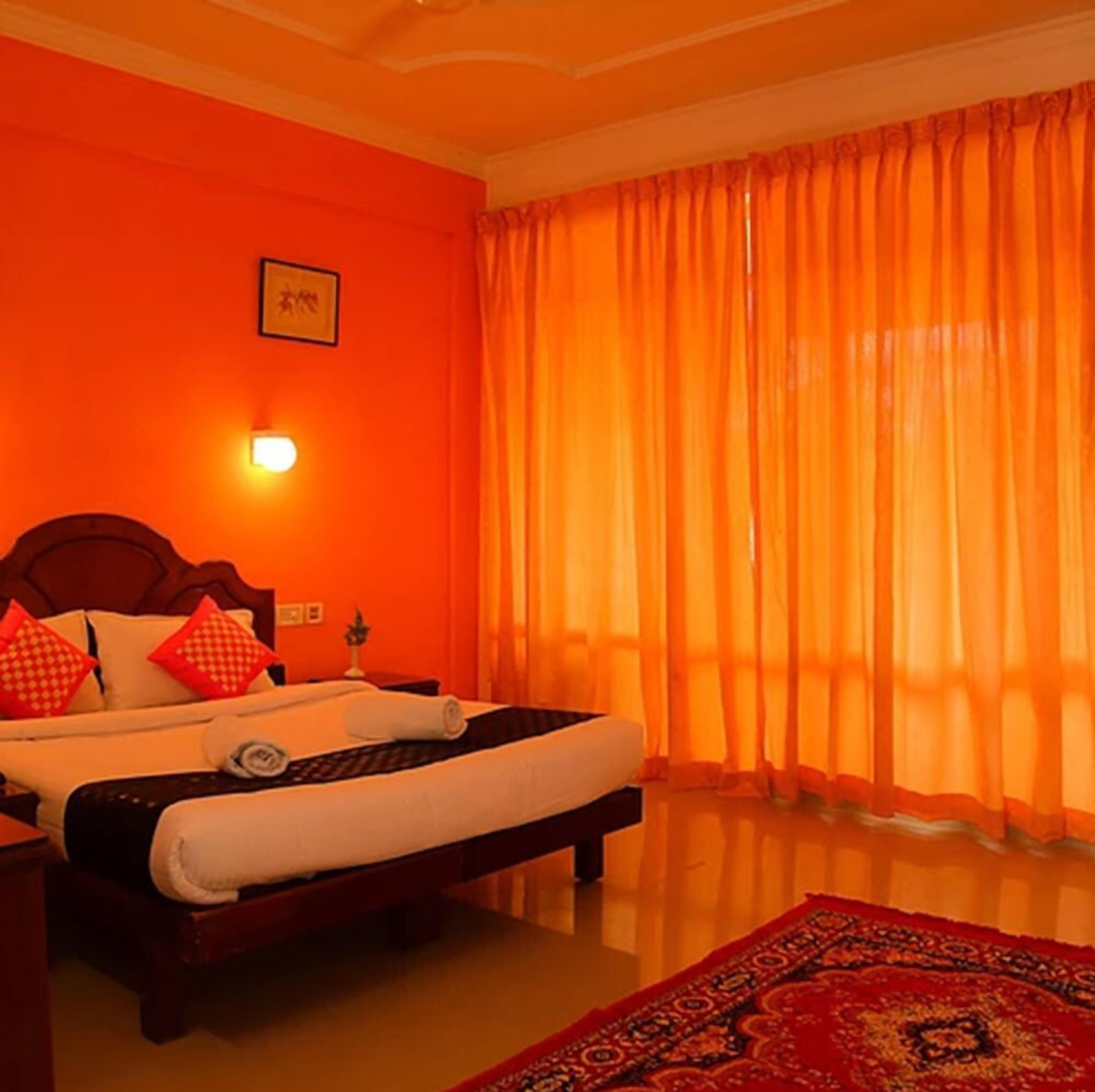 Een hotelkamer met een bed, nachtkastje, een wandlamp en een raam met oranje gordijnen.