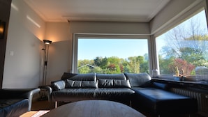 Un salon moderne avec un canapé d’angle, une lampe sur pied et une grande fenêtre donnant sur des arbres et une maison.