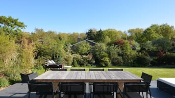 En uteplats med ett träbord och stolar, omgiven av frodig grönska och en klarblå himmel.