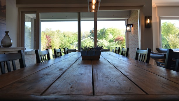 Une table à manger en bois, des chaises, un luminaire suspendu et une vue sur des arbres et une maison, le tout visible à travers de grandes fenêtres.