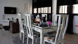 Una sala de estar moderna con una mesa de comedor de vidrio, sillas de madera, un televisor de pantalla plana y un ventanal con cortinas negras.