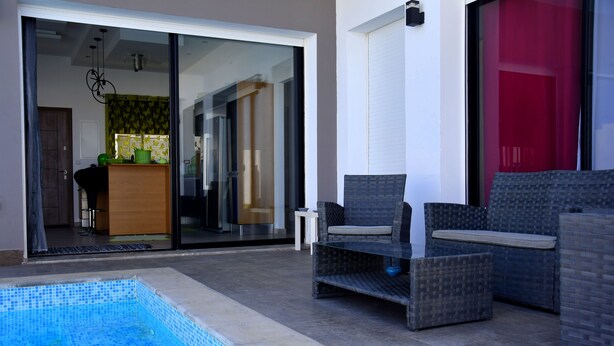 Un espacio exterior moderno con piscina, mobiliario de mimbre y un ventanal corredero que da a una cocina.