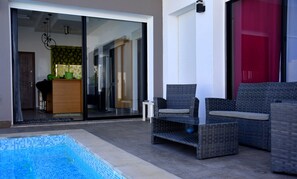Uma área externa moderna com piscina, móveis de vime e uma porta de vidro deslizante que dá para uma cozinha.
