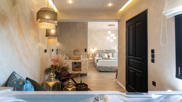 A modern hotel room with a large bathtub, a cozy sitting area with a sofa and decorative pillows, and a separate bedroom with a bed and a bathroom. - Archetypo (Naxos)