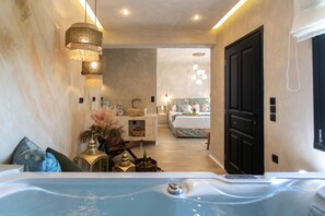 A modern hotel room with a large bathtub, a cozy sitting area with a sofa and decorative pillows, and a separate bedroom with a bed and a bathroom. - Archetypo (Naxos)