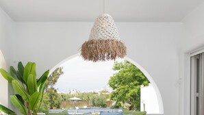 A modern living room with a large window, a sofa with colorful cushions, a wooden coffee table, and a hanging light fixture. - Archetypo (Naxos)