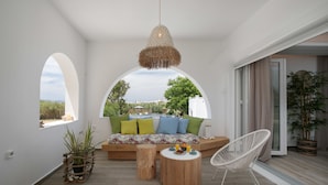 A modern living room with a sofa, a round wooden table, and a white chair. There is a hanging light fixture above the sofa and a potted plant on the floor. - Archetypo (Naxos)