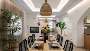 A dining area with a long table set for six, surrounded by black chairs, a large potted plant, and a hanging woven light fixture. - Archetypo (Naxos)