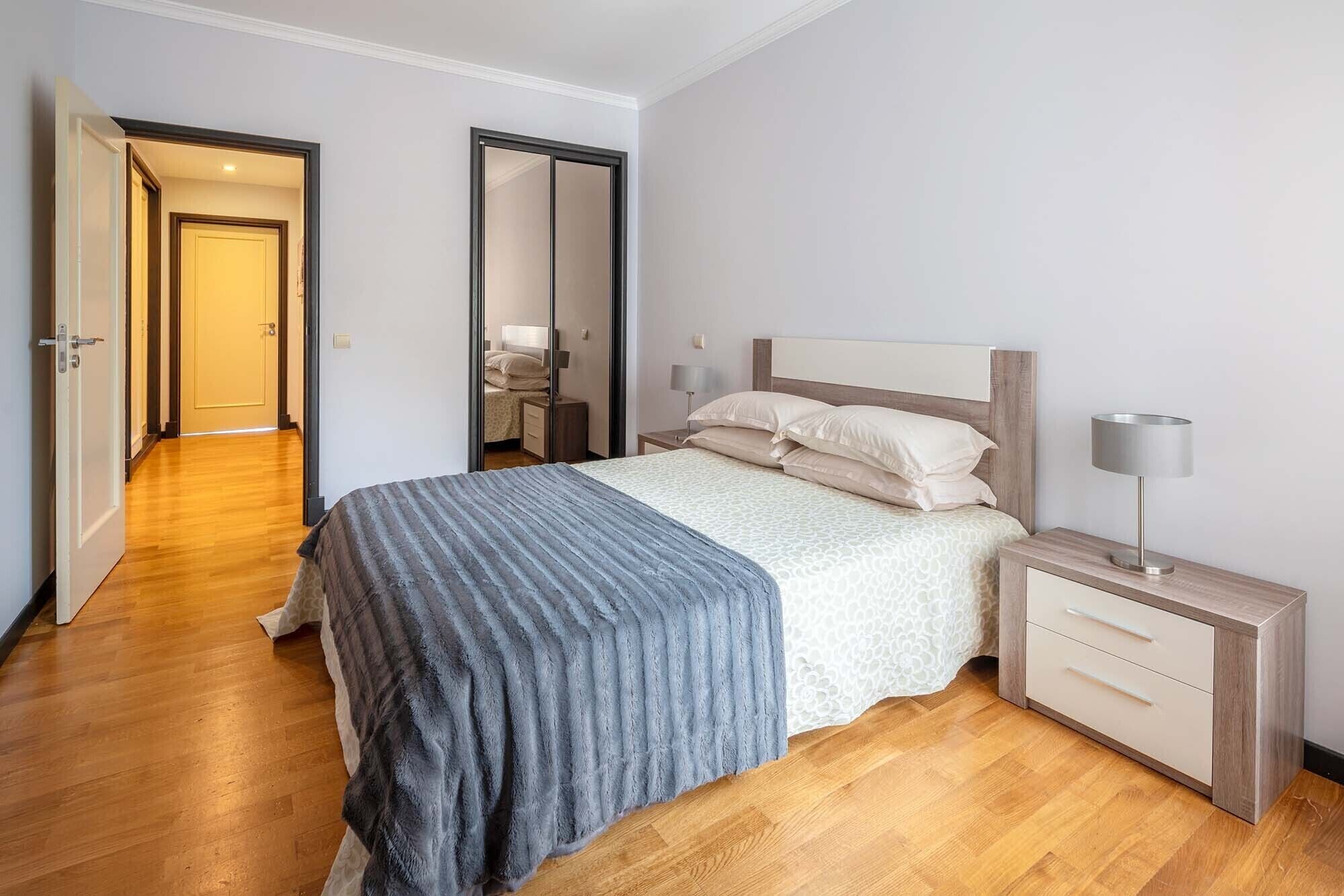 A bedroom with a bed, bedside tables, a mirror, and a door leading to another room.