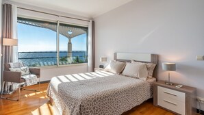 Una camera da letto con un'ampia finestra vista mare, un letto con copriletto decorato, un comodino con una lampada e una poltrona con cuscino.