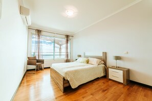 A bedroom with a bed, bedside table, lamp, chair, and a window with curtains. - Oceanside Terrace Apartment by Holiday Rental Management (Funchal)