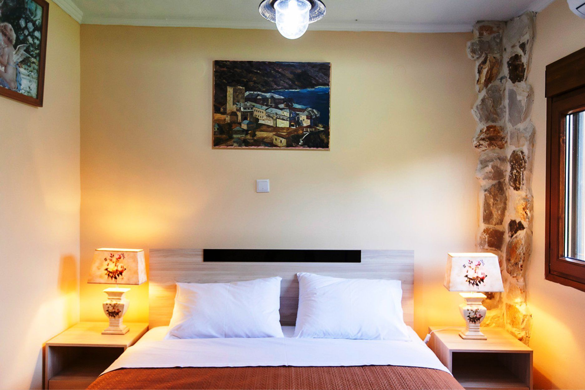 A hotel room with a bed, two bedside lamps, a painting on the wall, and a stone wall.