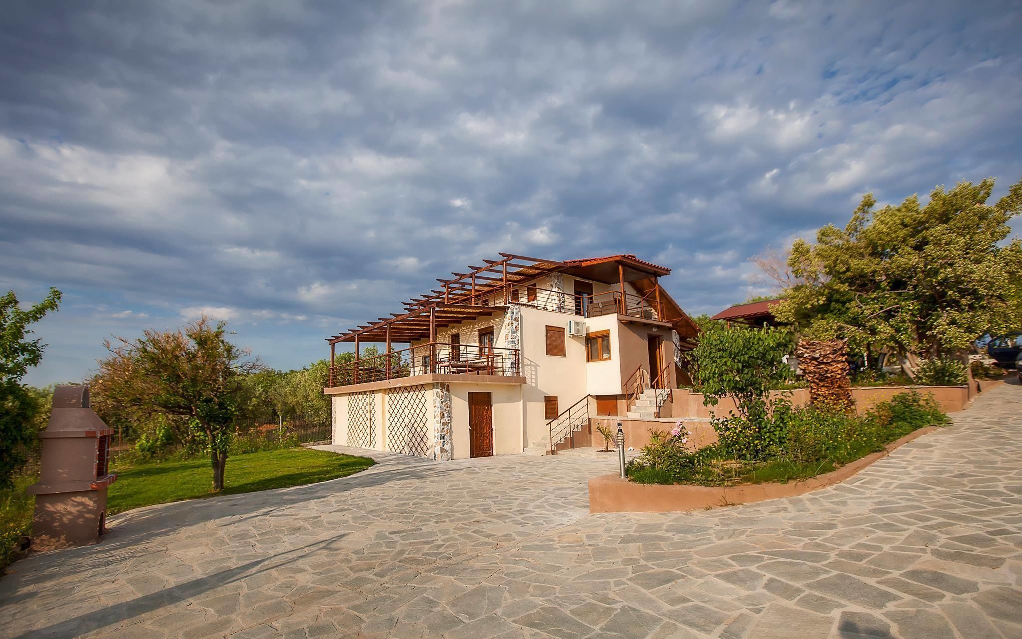 A two-story house with a stone-paved driveway, a wooden pergola, and a garden with various plants.