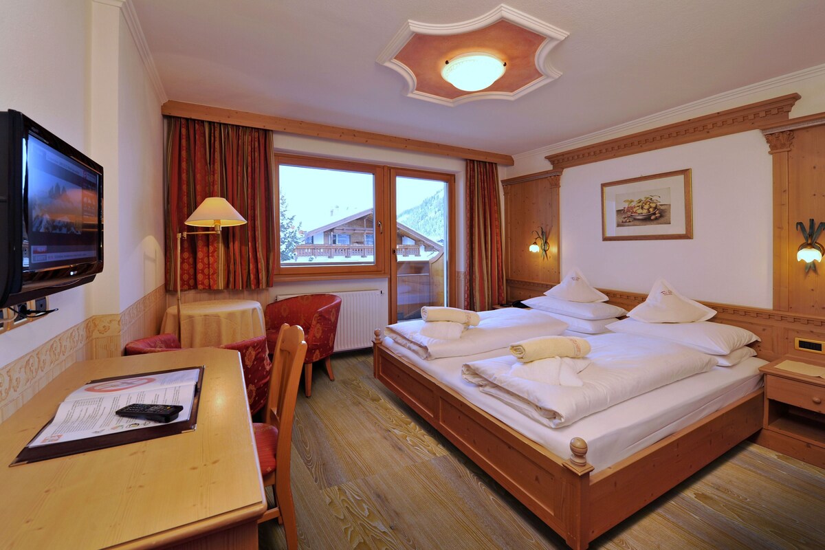 A hotel room with a wooden bed, a desk with a remote control, a chair, a table, a window with curtains, and a television on the wall.