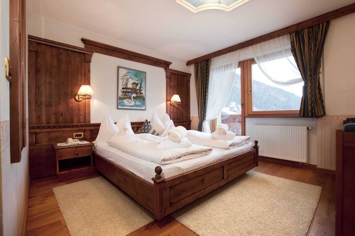 Un lit à baldaquin en bois, recouvert de draps blancs, avec deux lampes de chevet, un tableau au mur et une fenêtre avec des rideaux.