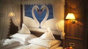 Une chambre d’hôtel avec un lit, deux oreillers, une table de chevet avec des pommes, et une décoration murale représentant des cygnes formant un cœur.