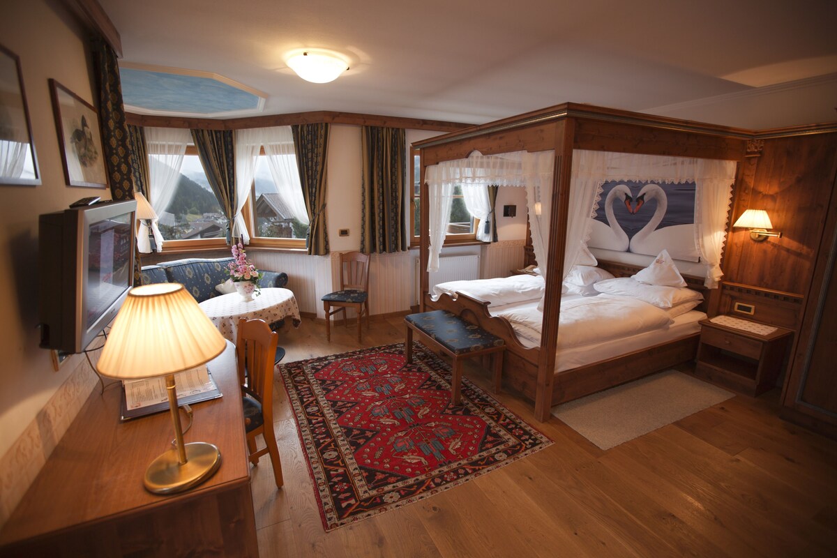 A four-poster bed with a heart-shaped window view, a wooden desk with a lamp, a small table with a vase, and a red patterned rug.